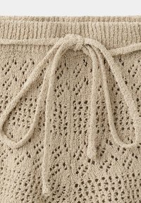 Tissu tricoté beige avec une ceinture côtelée et un nœud de cordon noué, présentant un motif losange ajouré semblable à de la dentelle.