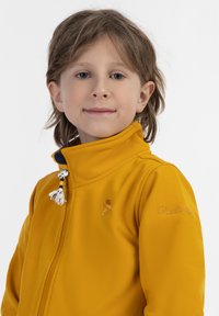 Den gule softshell-jacket har en høj krave, lynlås foran og marineblå detaljer med dekorative frynser og et broderet logo på ærmet.