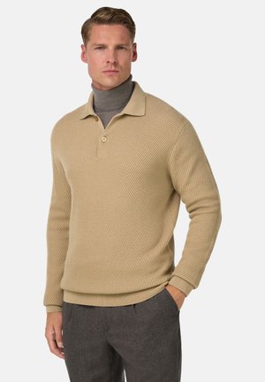 Pull en tricot beige avec une texture côtelée, doté d'un col et d'une fermeture à boutons, et de manches longues ; associé à un pantalon sombre.