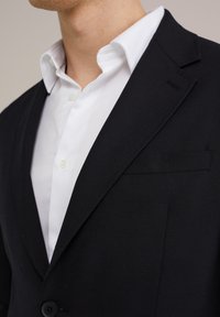 Must klasikaline ja musta tekstuuriga bleiser, millel on nurgelised kraed, ühe nööbi kinnitamine ja vasakul rinnatasku. Allpool on valge kraedega särk.