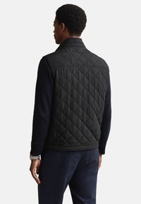 Zwarte gewatteerde bodywarmer met een hoge kraag en elastische zoom, met een gladde textuur en ruitpatroon, gedragen over een donkere trui.