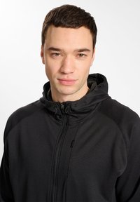 Hanorac negru cu fermoar, cu guler înalt, dintr-un material texturat și cu sireturi ajustabile. Are un fermoar frontal neted și panouri laterale.