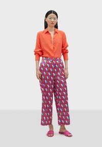 En orange knäpp skjorta, mönstrade flerfärgade byxor med vida ben och geometrisk design, samt rosa platta sandaler, mot en neutral bakgrund.