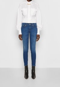 Vit skjorta med krage och två framfickor, kombinerad med blå tajta jeans. Modellen bär svarta ankelboots. Enkelt mönster, släta texturer.