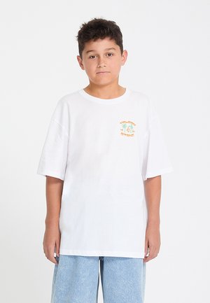 Garçon aux cheveux courts et foncés portant un t-shirt blanc oversize avec un petit motif coloré sur la poitrine et un short en jean bleu clair.