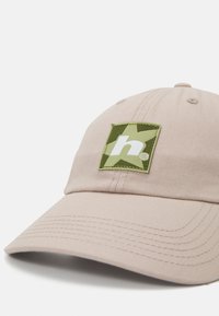 Beige bomullslue med buet skjerm, med en grønn stjerneapplikasjon med et stilisert 'h'-symbol. Strukturert stoff med innsydd detaljer.