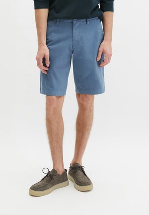 Man staat en draagt blauwe knielange shorts, een donkergroene trui en grijze suède veterschoenen met beige zolen.