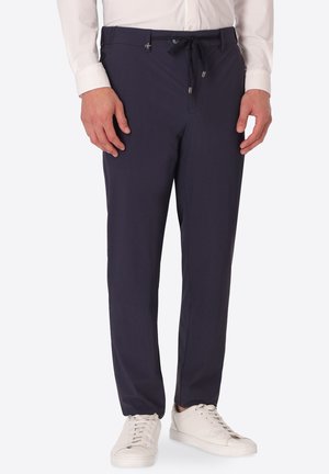 Uomo con pantaloni blu scuro con coulisse, scarpe da ginnastica bianche e camicia bianca a maniche lunghe, in piedi davanti a uno sfondo semplice.