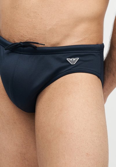 Κοντινό πλάνο ενός ατόμου που φοράει μαύρα μαγιό με κορδόνι και λευκό λογότυπο στο αριστερό ισχίο.