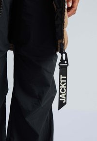 Zwart oversized buitenkleding met een beige capuchon, voorzien van een opvallend logo-label met de tekst "JACK1T" en een zwarte ritssluiting. De stof lijkt glad te zijn.