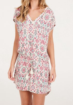 Kobieta w sukience z krótkim rękawem i dekoltem w serek, ozdobionej różowo-niebiesko-szarymi geometrycznymi wzorami oraz ściągaczem w talii z frędzlami.