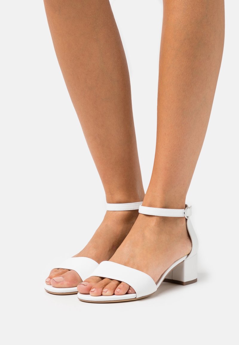 Pieds portant des sandales blanches à talon bloc ouvert aux orteils avec brides à la cheville sur un fond clair uni.