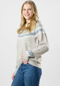 Lächelnde Frau mit langen blonden Haaren, die einen beigen gemusterten Pullover und blaue Jeans trägt und mit einer Hand in ihrer Tasche steht.