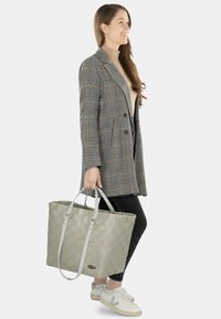 Gewebte graue Tragetasche mit hellgrauen Tragegriffen, rechteckiger Form und glatter Textur, kombiniert mit einem karierten Blazer und schwarzen Hosen.