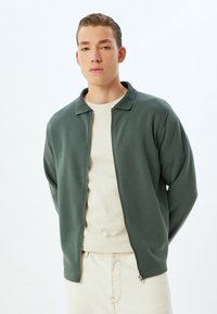 Chaqueta verde con cremallera y cuello acanalado, hecha de una tela suave, llevada sobre un suéter de cuello redondo en un tono beige claro y combinada con pantalones claros.