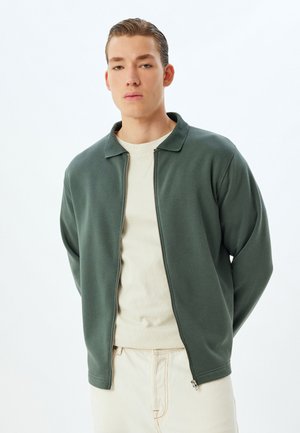 Chaqueta verde con cremallera y cuello acanalado, hecha de una tela suave, llevada sobre un suéter de cuello redondo en un tono beige claro y combinada con pantalones claros.