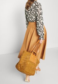 Borsa a rete in paglia arancione con manici in cuoio, forma quadrata, e un pannello solido con il nome del marchio, abbinata a una gonna beige.