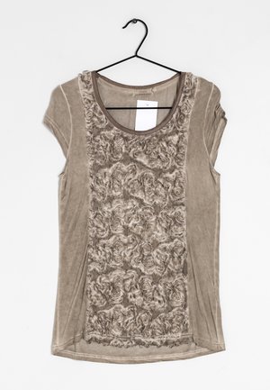 Beige overhemd met korte mouwen met een gestructureerd bloemmotief aan de voorkant, ronde hals, hangend aan een zwarte draadhanger met een wit label aan de binnenkant van de kraag.