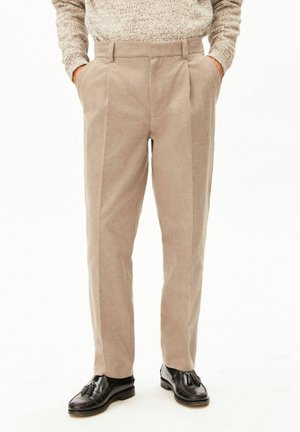 Beige Cordhosen, getragen mit den Händen in den Taschen und kombiniert mit schwarzen Leder-Tassel-Loafern vor weißem Hintergrund.