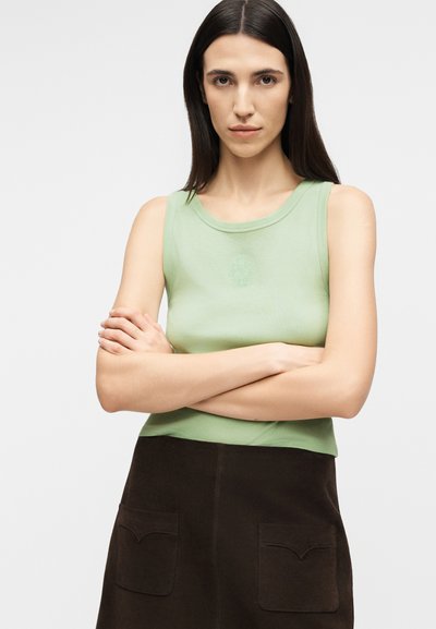 Νεαρή γυναίκα με ίσια σκούρα μαλλιά, φοράει ανοιχτό πράσινο αμάνικο τοπ και σκούρο φούστα με μπροστινές τσέπες, με σταυρωμένα χέρια και ουδέτερη έκφραση.