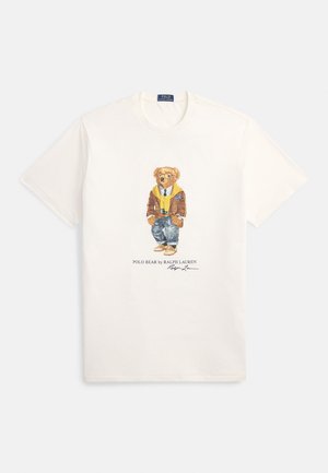 Hvit bomulls T-skjorte med et motiv av en kledd bjørn i brun jakke, gul genser og blå jeans; "Polo Bear by Ralph Lauren" trykt under.
