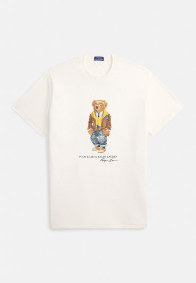 Bela bombažna majica z grafiko oblečenega medveda v rjavi jakni, rumenem puloverju in modrih kavbojkah; spodaj je natisnjen napis "Polo Bear by Ralph Lauren".