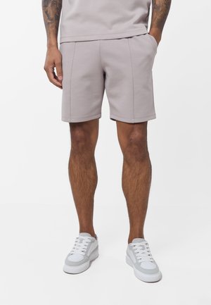Mann trägt hellgraue Shorts und weiße Sneakers, steht mit den Händen in den Taschen vor einem einfarbigen Hintergrund.