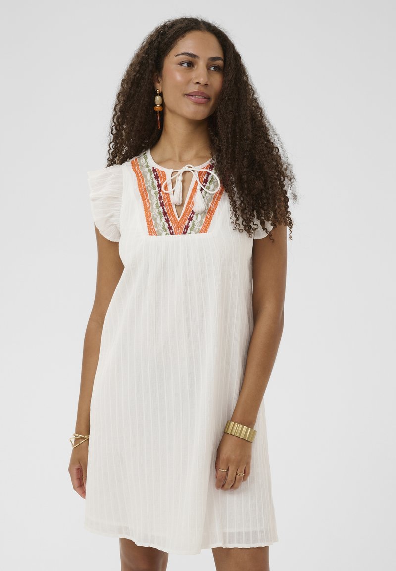 Femme aux cheveux bouclés portant une robe blanche avec des détails brodés orange et verts sur le col, accessoirisée avec des boucles d'oreilles et des bracelets.