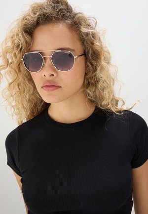 Donna con capelli biondi ricci che indossa una maglietta nera a costine con maniche corte e occhiali da sole sfumati con montatura dorata, che guarda dritto davanti a sé con espressione neutra.
