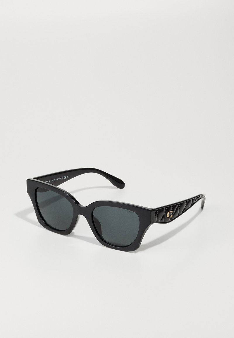 Lunettes de soleil noires avec des montures angulaires et des verres sombres. Branche texturée noire avec un motif matelassé et un logo doré en accent sur le côté.