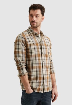 Homme barbu portant une chemise à manches longues à carreaux marron et beige et un jean foncé, mains dans les poches, regardant sur le côté sur fond blanc.