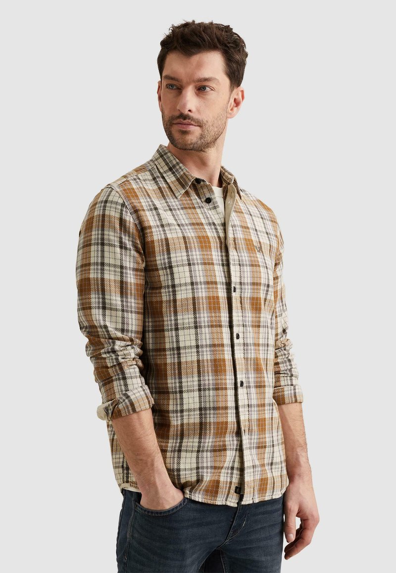 Hombre con barba que lleva una camisa de manga larga a cuadros marrón y beige y vaqueros oscuros, con las manos en los bolsillos, mirando hacia un lado contra un fondo blanco.