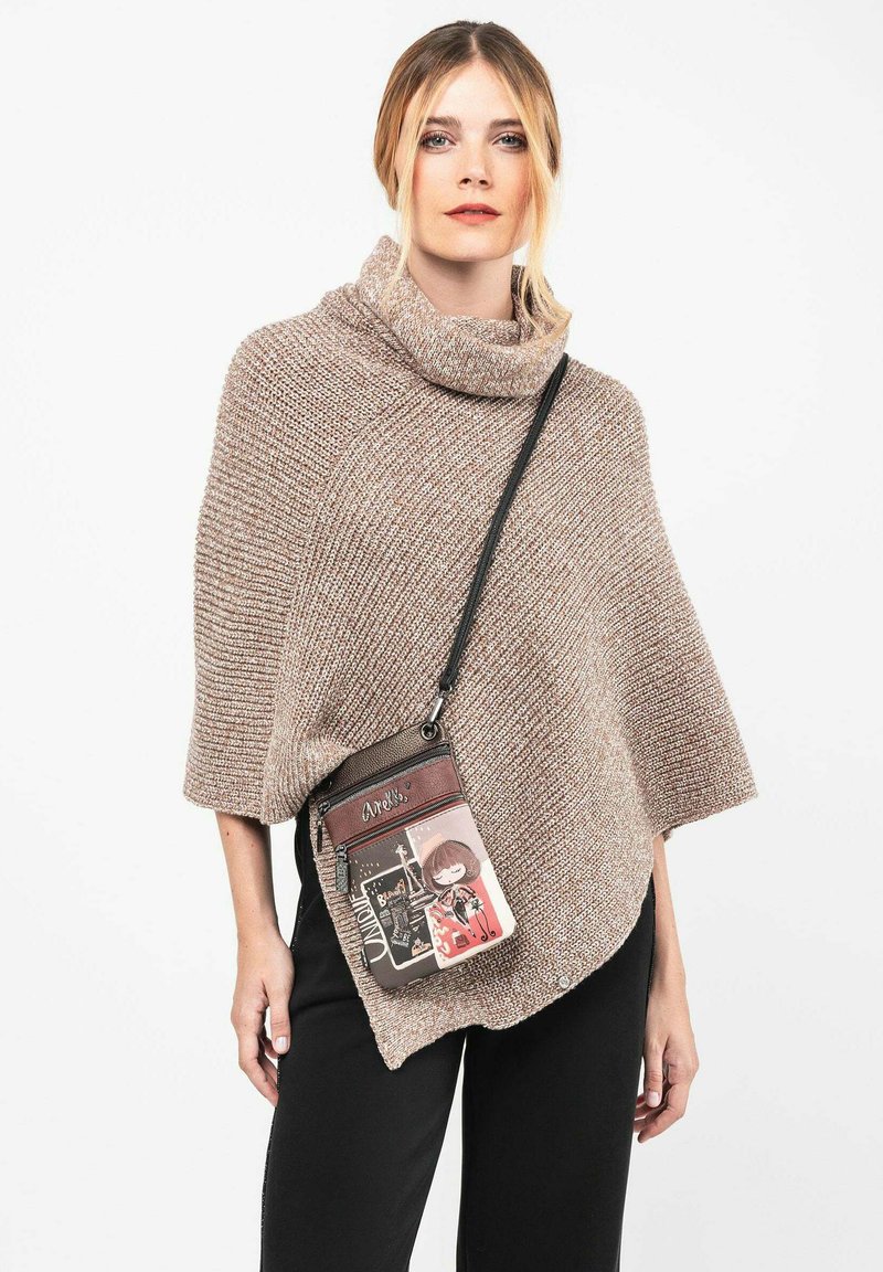 Beige gestrickter Poncho mit hohem Kragen und lockerer Passform. Kombiniert mit schwarzen Hosen und einer kleinen, gemusterten Umhängetasche.