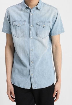 Mężczyzna ubrany w jasnoniebieską, krótkorękawową koszulę dżinsową z zapiętymi kieszeniami na piersi i czarne spodnie na jednolitym tle.