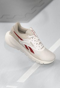 Zapatilla deportiva beige con una superficie texturizada, acentos rojos, cordones blancos y una suela acolchada. Presenta un logo de la marca en el lateral.