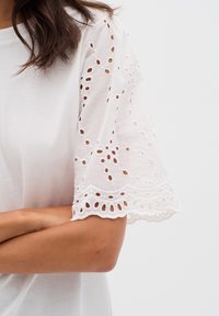 T-shirt bianco a maniche corte con raffinata ricamatura a occhio e orlo scallop. Texture del tessuto liscia, semplice scollo rotondo.