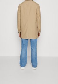 Tan waterdichte jas met een rechte snit, knoopmanchetten en een rugventilatie, gecombineerd met lichtblauwe flare jeans en witte sneakers.