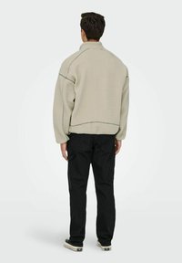 Giacca in pile color beige chiaro con colletto alto e cuciture a contrasto, abbinata a pantaloni neri larghi e sneakers.