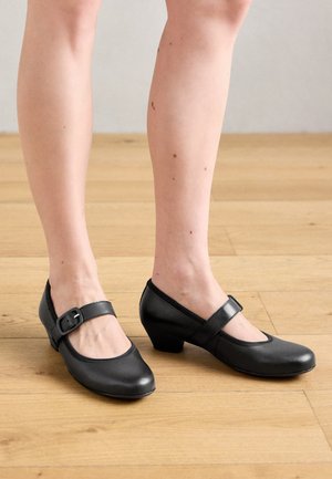 Osoba koja nosi crne kožne Mary Jane cipele s niskim potpeticama na drvenom podu, prikazano obaju stopala i donjih nogu.