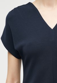 Γυναίκα που φοράει μπλούζα με V λαιμόκοψη και κοντά μανίκια σε σκούρο μπλε χρώμα, εμφανίζοντας το πάνω μέρος του κορμού και ένα μέρος του λαιμού και του χεριού, με φόντο απλό.