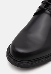 Chaussure en cuir noir avec une finition lisse et brillante, à bout rond. Semelle en caoutchouc avec des détails en arête. Design minimaliste, sans marquage visible.