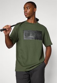 Olivengrønn kortermet t-skjorte med en strukturert svart logopatch som har sports emblemer. Glatt stoff og avslappet passform.