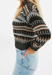 Schwarzer Strickpullover mit geometrischen Mustern in Weiß, Beige und Grau. Hat einen hohen Kragen und gerippte Bündchen, kombiniert mit hellblauen Jeans.