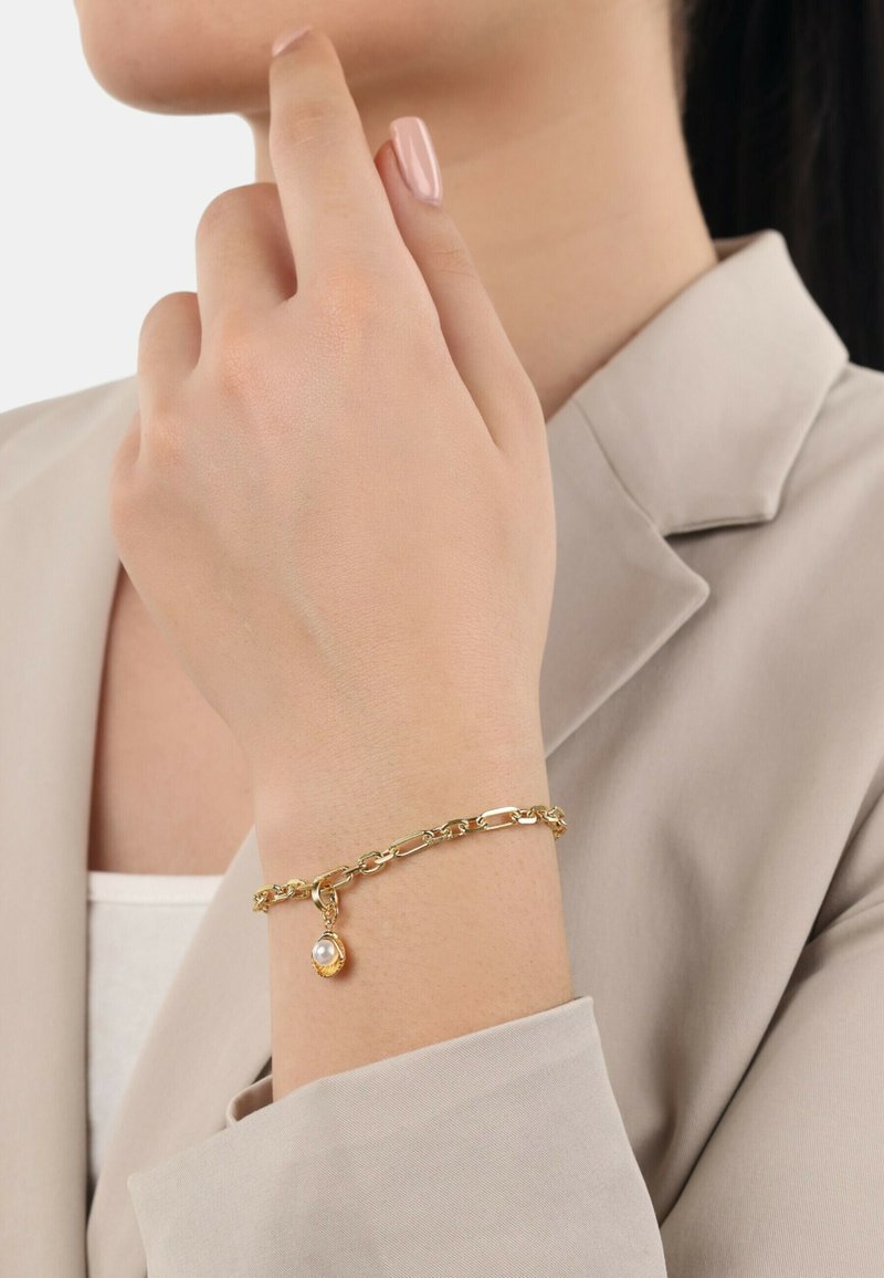 Bracciale in oro con charm a forma di perla, che mostra un design intrecciato e una superficie liscia su una manica di colore chiaro.