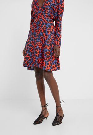 Frau in einem knielangen Kleid mit rotem, blauem und schwarzem Muster und schwarzen, schnürbaren, niedrig-absätzigen Schuhen, die vor einem weißen Hintergrund steht.