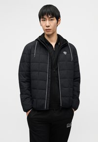 Μαύρο μπουφάν με επένδυση και σχεδιασμό με ραφές, με κουκούλα και ασημένιες λεπτομέρειες. Φορεμένο πάνω από μαύρο μπλουζάκι με φερμουάρ και μαύρη φόρμα.