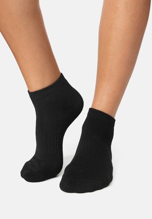 Para stóp w czarnych stopkach do kostki na białym tle, jedna stopa lekko uniesiona, aby pokazać teksturę i dopasowanie skarpetki.