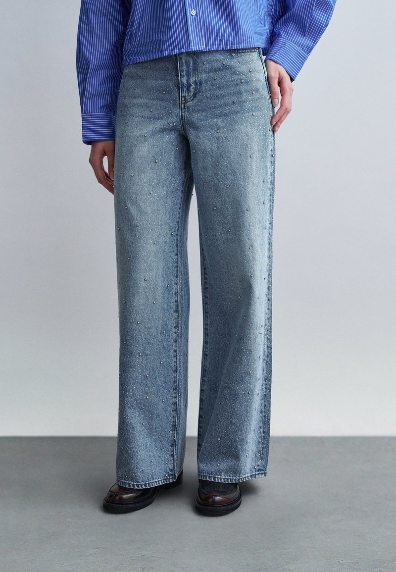 Άτομο που φοράει ανοιχτό γαλάζιο τζιν με φαρδιά μπατζάκια και μικρές διακοσμητικές πέρλες, σκούρα παπούτσια και μπλε ριγέ πουκάμισο με κουμπιά.