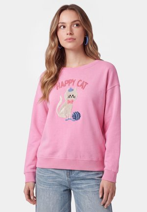 Giovane donna che indossa una felpa rosa con una grafica di un gatto e un gomitolo di lana con le parole "Happy Cat" sopra, abbinata a jeans azzurri.