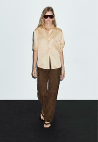 Hellbeige Satin-Hemd mit Knöpfen, braune Wildlederhose, schwarze Sonnenbrille und flache Sandalen. Glatte Textur mit lockerer Passform.